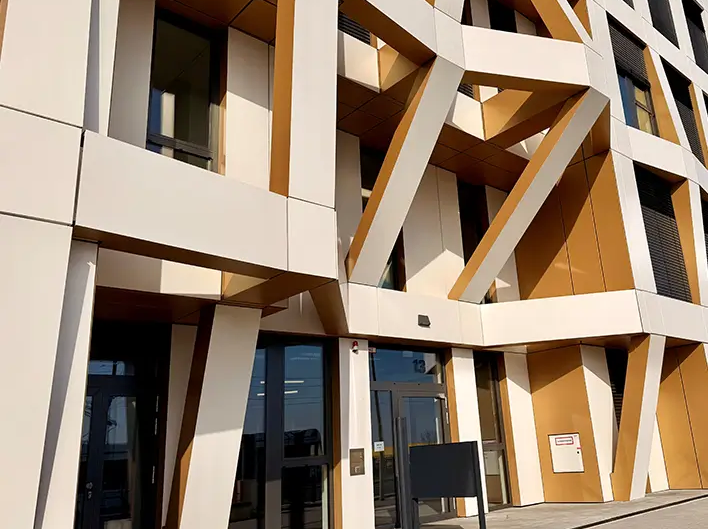 Szczegółowe ujęcie strefy wejściowej Brainfactory Bochum z charakterystyczną strukturą elewacji, ukośnymi elementami wsporczymi oraz nowoczesną architekturą w białych i złotych tonacjach.