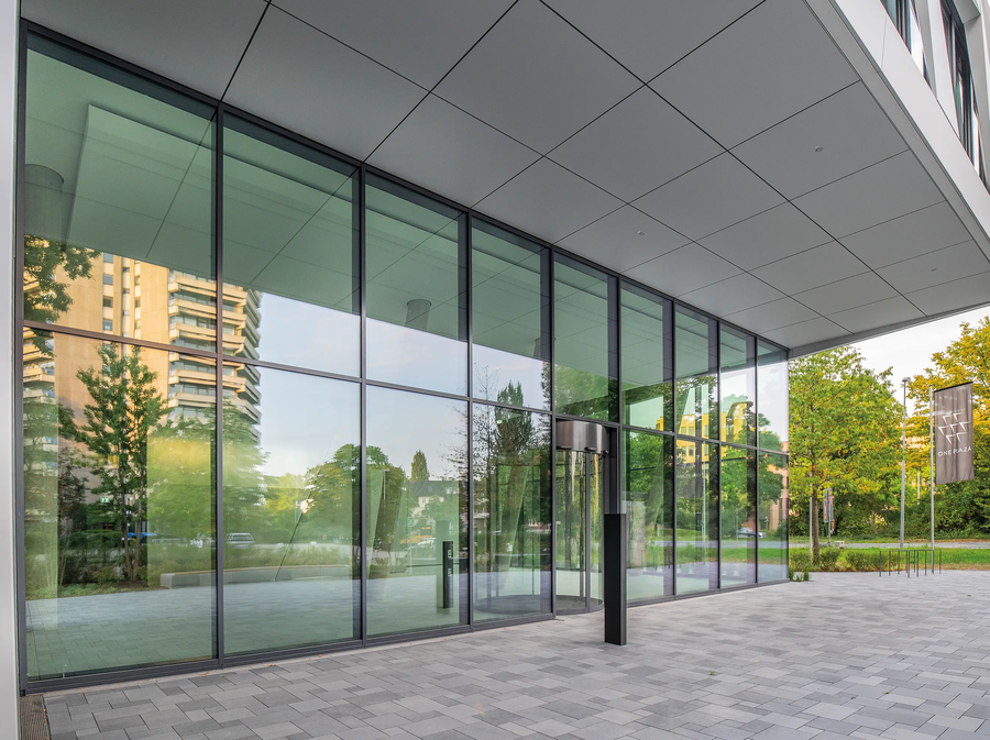 Wejście do ONE PLAZA z przestronną szklaną fasadą, nowoczesną architekturą i widokiem na okoliczne budynki oraz tereny zielone.